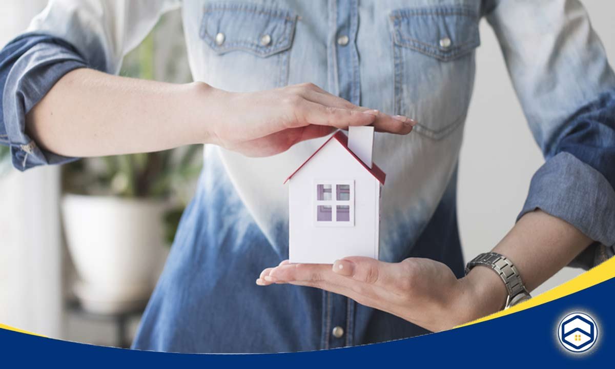 A person holds a small model house with both hands, creating a protective gesture above it