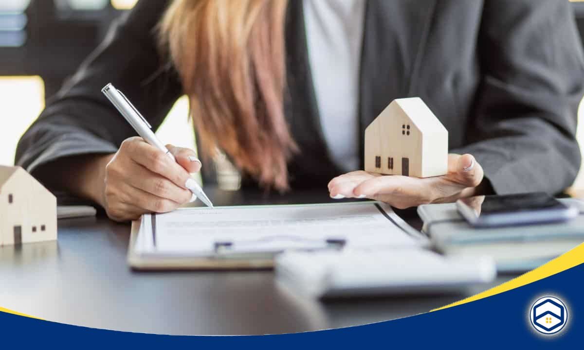 A woman in a suit writes on a document while holding a small wooden house model 