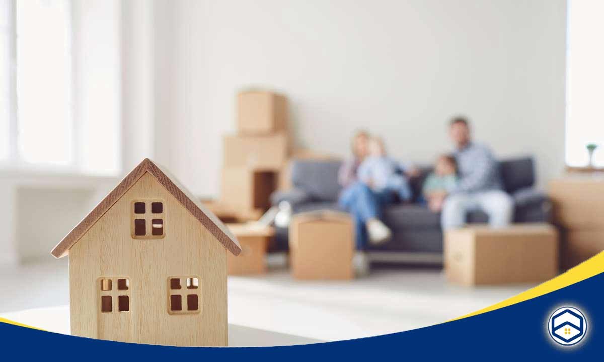 A wooden house model is in the foreground, with a blurred family sitting on a couch in the background, representing the context of renting versus buying a condo.