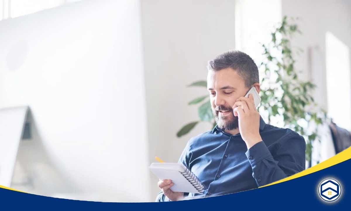 The image shows a man in a blue shirt talking on a phone, with a laptop and plant in the background.