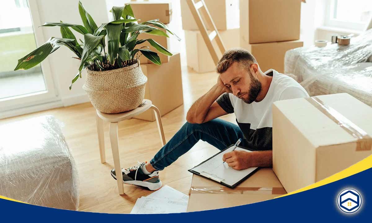 The image shows a man sitting on the floor of a room surrounded by moving boxes, appearing to be planning or organizing his move.