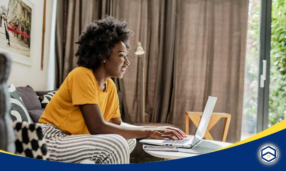 The image shows a young woman with curly hair sitting at a desk and using a laptop computer. She appears to be in a home setting, with curtains