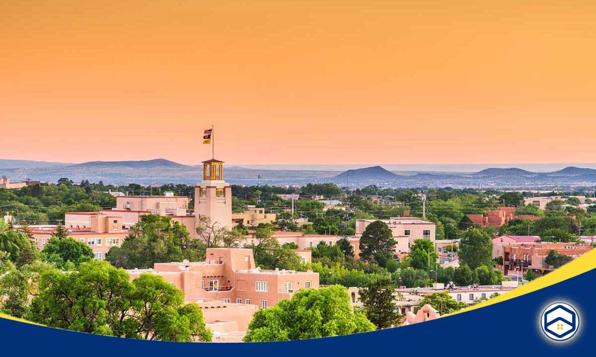 The image shows a panoramic view of the historic city of Santa Fe, New Mexico, with its distinctive adobe architecture and surrounding mountains.