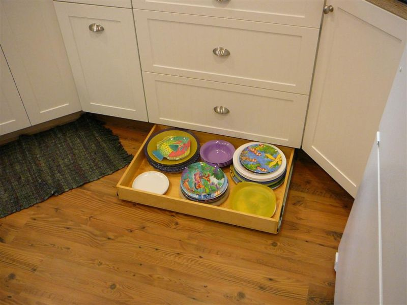 A pull-out drawer installed in a lower kitchen cabinet, filled with colorful plates of various sizes. The white cabinets contrast with the wooden floor, highlighting an ergonomic and space-saving kitchen organization idea.
