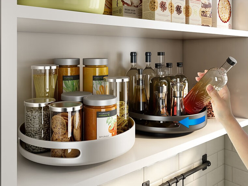 A Lazy Susans rack holding oil bottles and spices, with a hand reaching for a bottle. Next to it is another rack with jars of dry food.