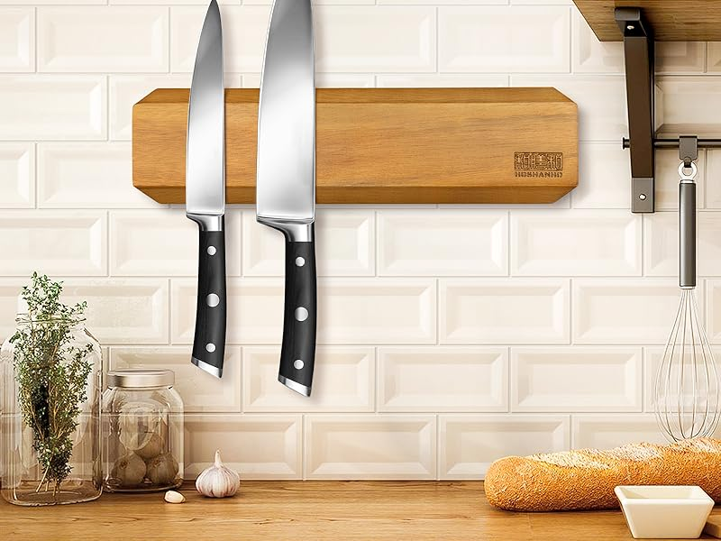 A wooden magnetic knife strip mounted on a white subway tile wall, holding two black-handled knives, with a whisk and herbs nearby.