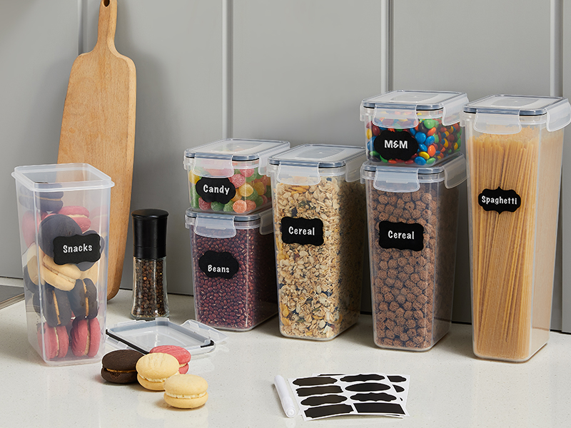 A collection of transparent plastic food storage containers with black labels, neatly filled with various dry foods like cereal, beans, pasta, and candy, arranged on a kitchen countertop.
