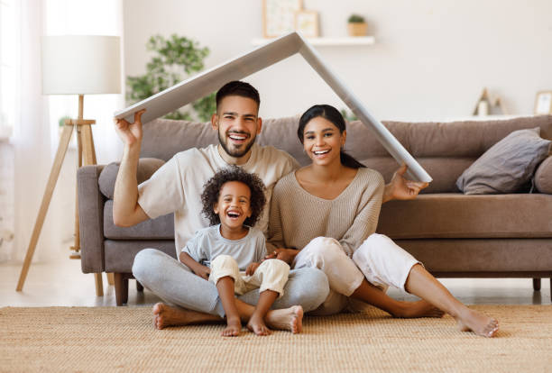 Minimalist Houston family sitting on the floor of a clean, cozy living room, smiling together while holding a roof-shaped panel symbolizing a peaceful, simplified lifestyle at home.