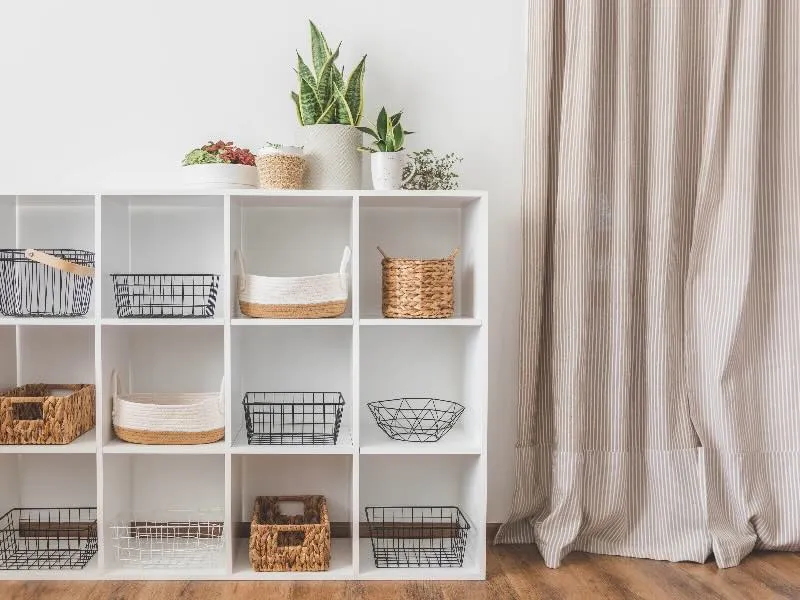 White cubby shelving unit featuring organized wire and woven baskets with houseplants on top, representing practical minimalist home organization in a Houston-style interior.