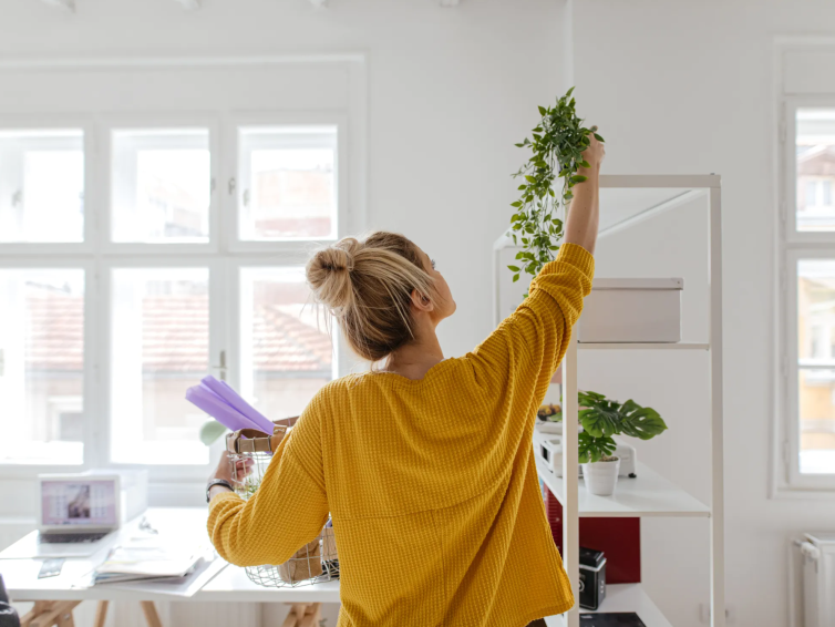 Woman in mustard sweater arranging greenery in a bright, minimalist home office in Houston, with clean white shelving and clutter-free surfaces for a calm, efficient lifestyle.