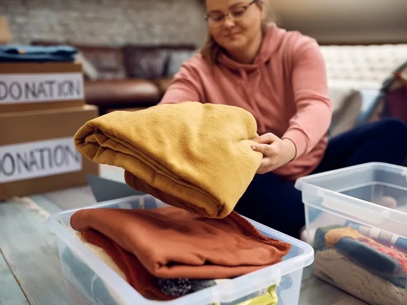 Woman decluttering her home by sorting out clothing for donation using clear plastic bins, part of a space optimization strategy to minimize clutter and organize small spaces more efficiently.