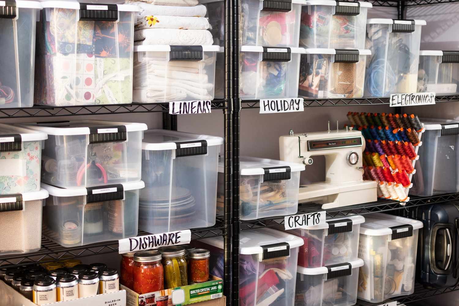Utility storage shelves filled with transparent labeled bins for crafts, dishware, electronics, linens, and holiday items. A sewing machine and preserved food jars emphasize practical home organization.