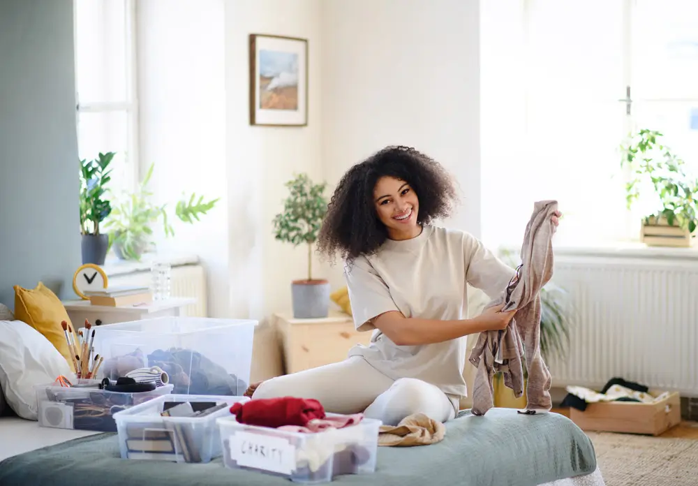Happy young woman folding and organizing clothes in a bright, minimalist bedroom, utilizing labeled storage bins for better space optimization and home organization.